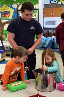 High school student standing next to 2 preschool students stirring a pot
